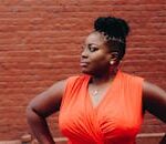 Portrait of a confident woman in a red dress posing against a brick wall.